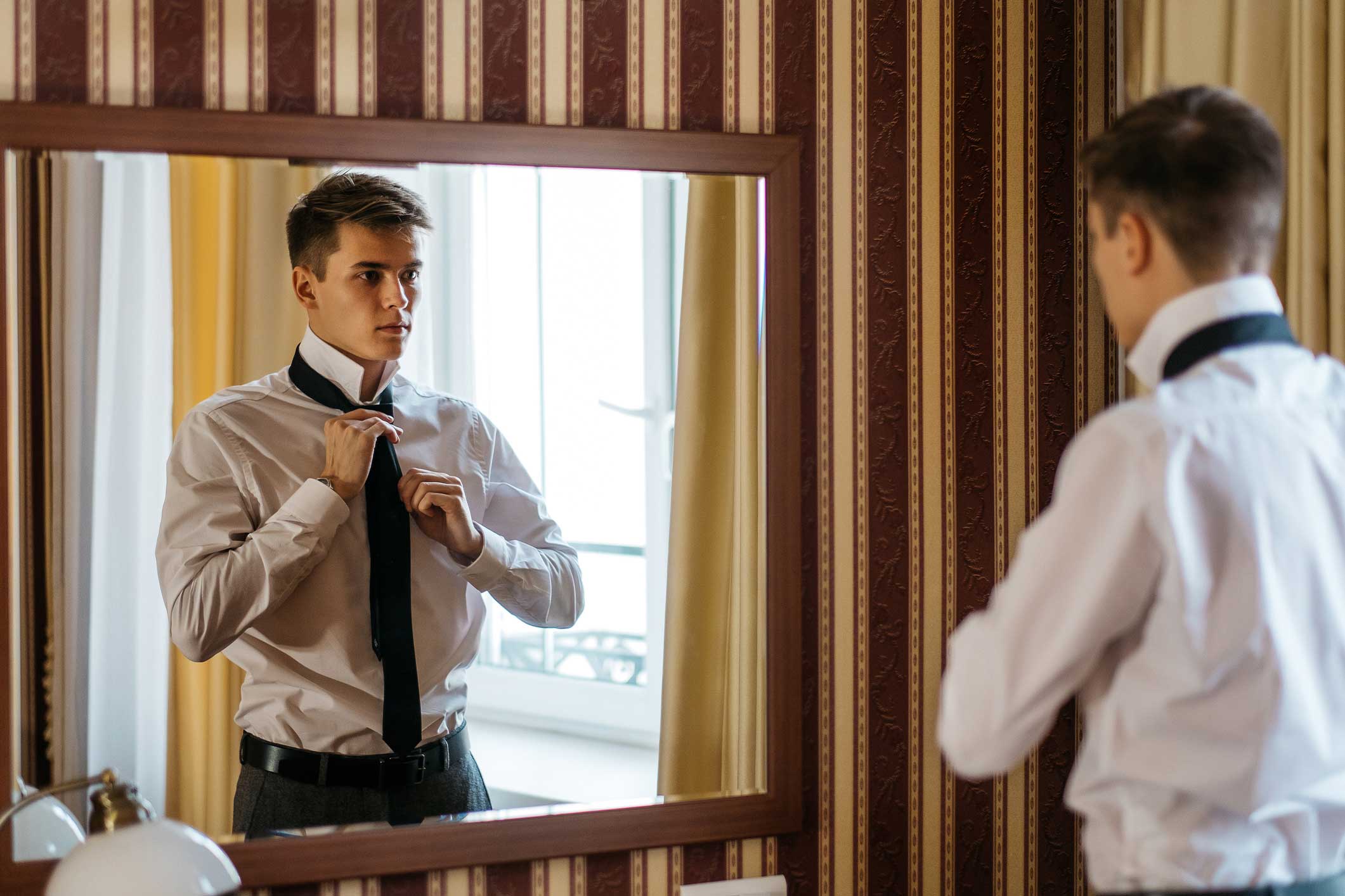A young man tying his tie over his shirt in a mirror